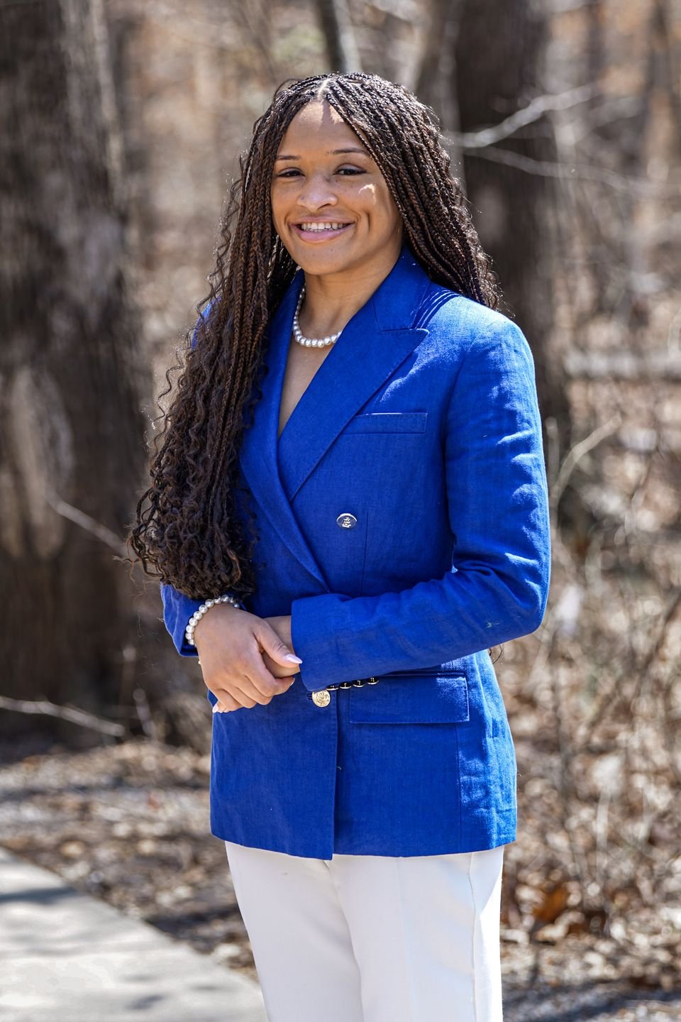 A smiling professional in a royal blue double-breasted blazer and white trousers, photographed outdoors among trees.