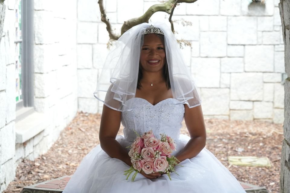 A bride in a strapless gown and veil, seated with a bouquet of pink roses, smiling.