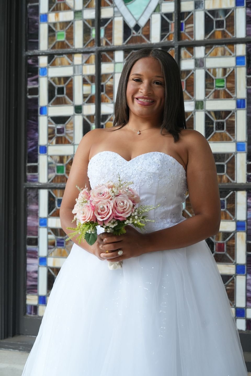 A bride in a strapless gown stands in front of a stained glass window holding pink roses.