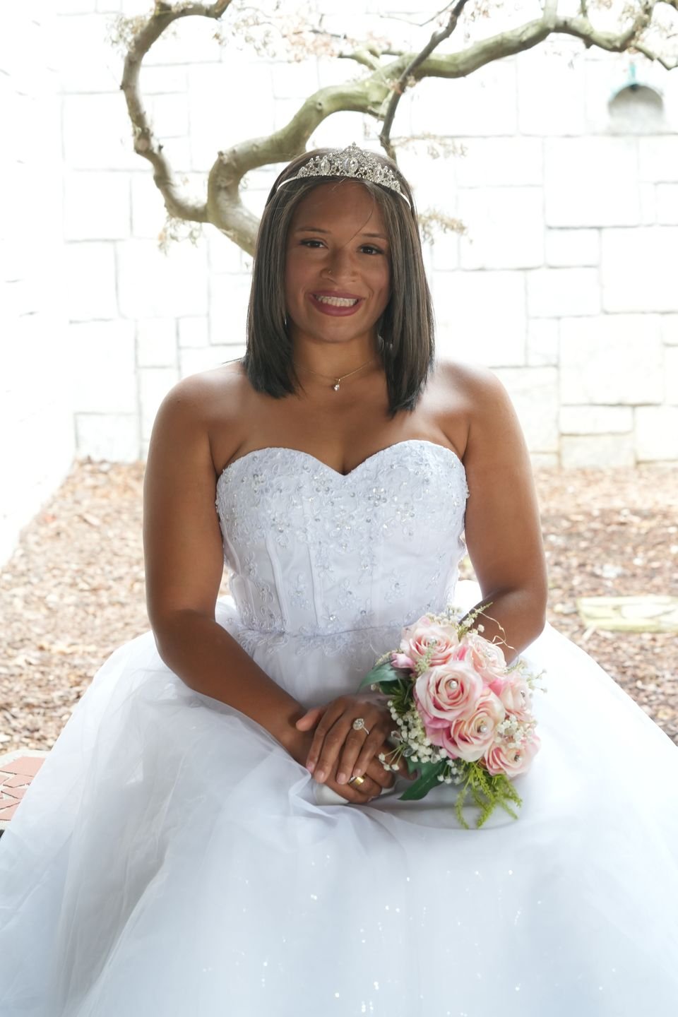 A bride in a strapless gown and tiara sits with a bouquet of pink roses.