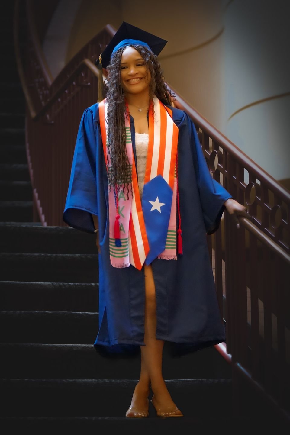 A graduate in a navy gown and flag stoles poses on a staircase.