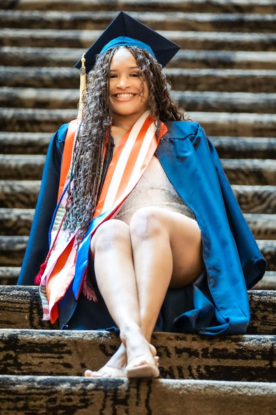 A graduate in a blue gown and stoles sits on outdoor stone steps, smiling.