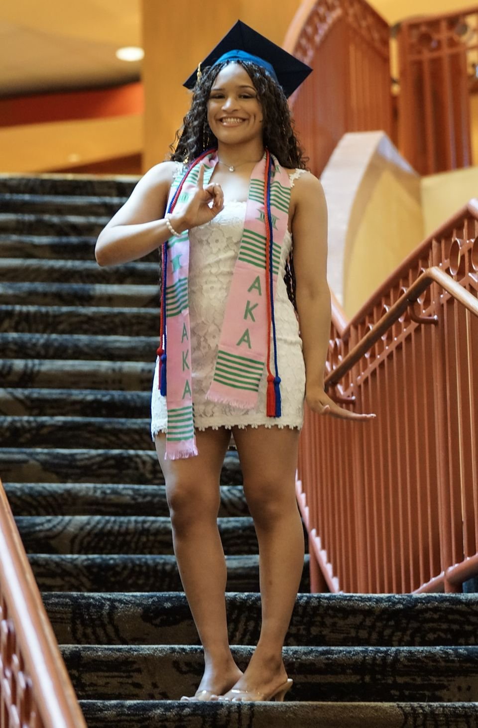 A graduate in a cap and a kente stole stands on a carpeted staircase.