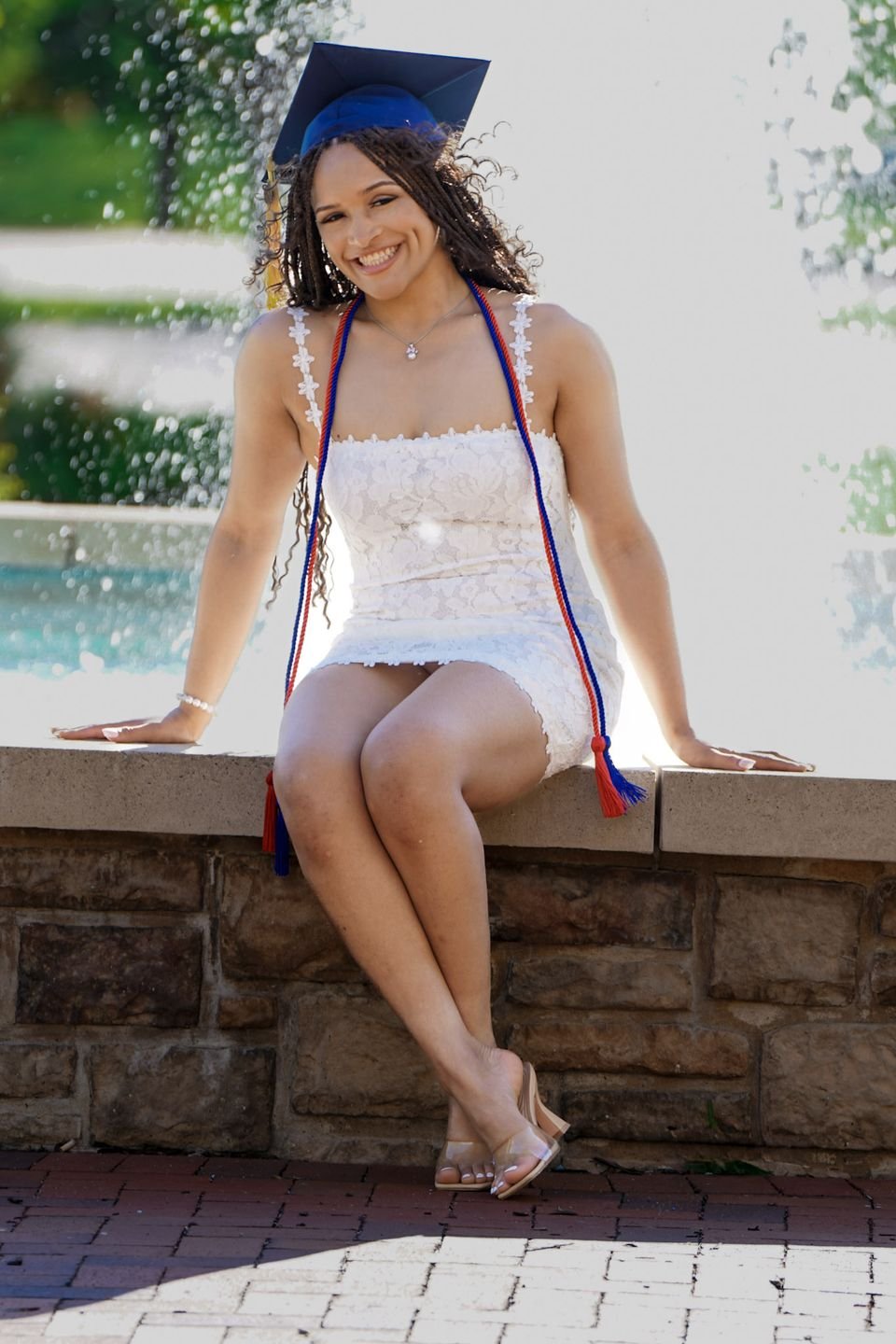 A graduate in a cap and white dress sits on a fountain wall with honor cords.