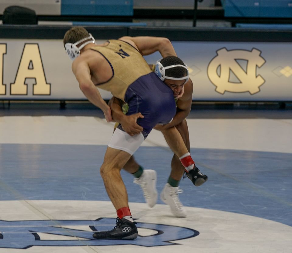 A wrestler in navy and gold works for a takedown on a blue collegiate mat.