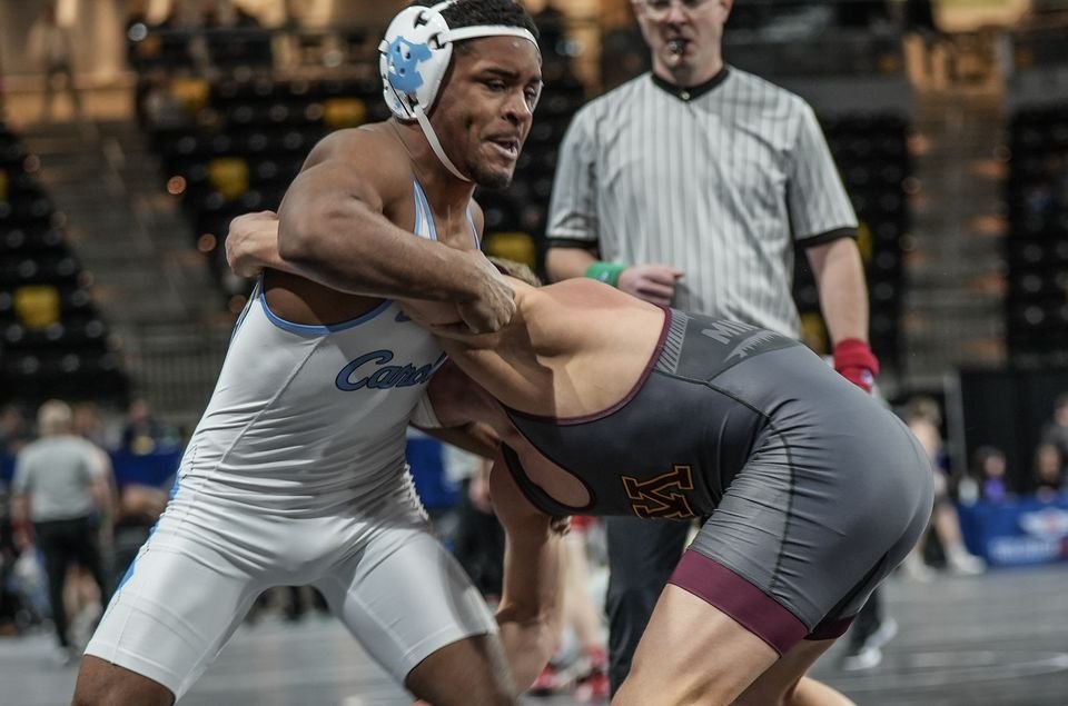 A wrestler in white controls his opponent's arm while a referee watches.