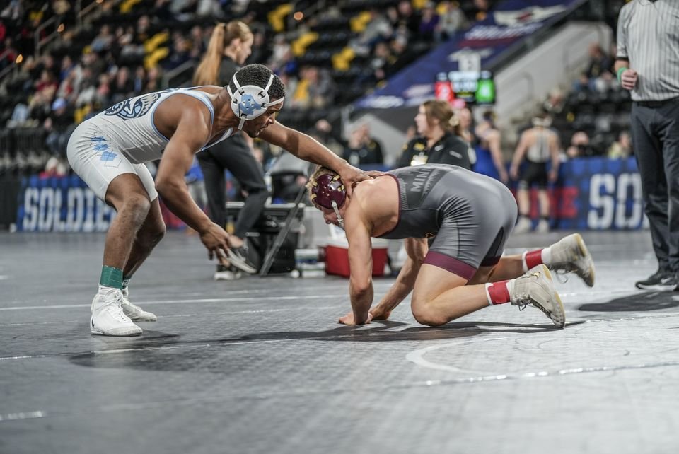 A wrestler in grey reaches for his opponent's head as they square off on a grey mat.