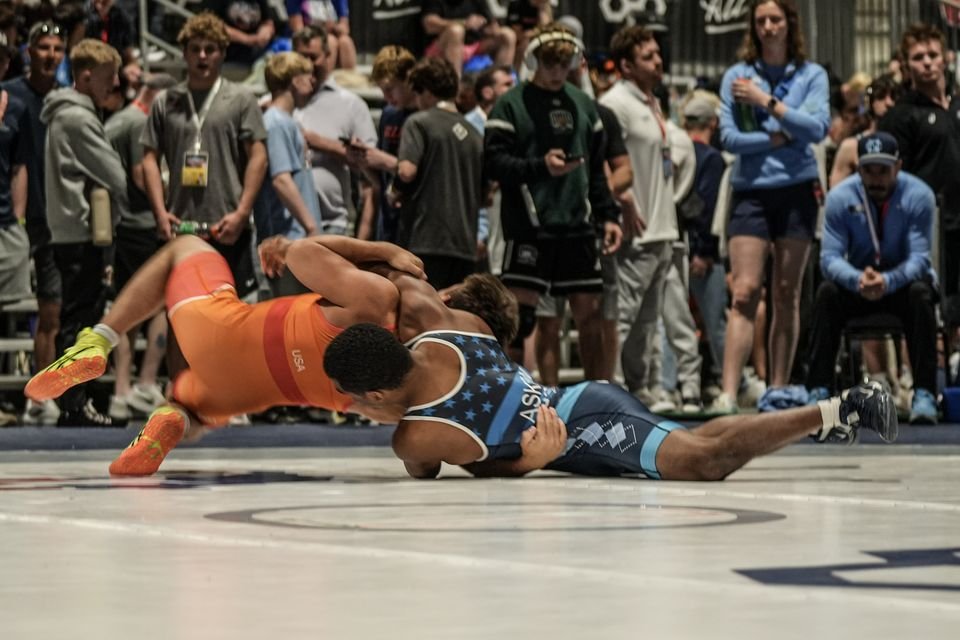 A wrestler in orange drives over his opponent in navy in front of a packed crowd.