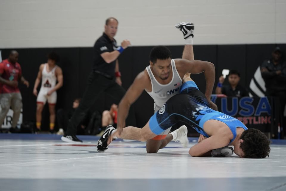 A wrestler in a white singlet drives his opponent in blue down to the mat.