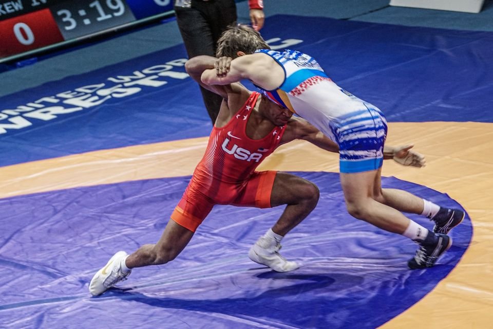 A wrestler in a red USA singlet drives low into his opponent in blue and white on an international mat.