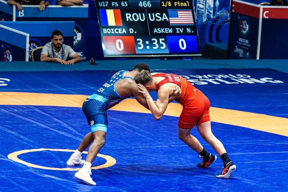 Two wrestlers, one in a USA singlet and one in red, lock up in a collar tie at a world championship match.