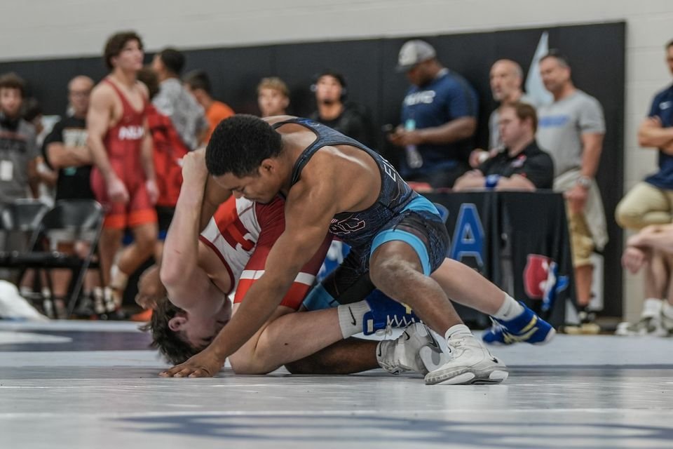 A wrestler in navy and light blue works over his opponent in red on a grey mat as spectators look on.