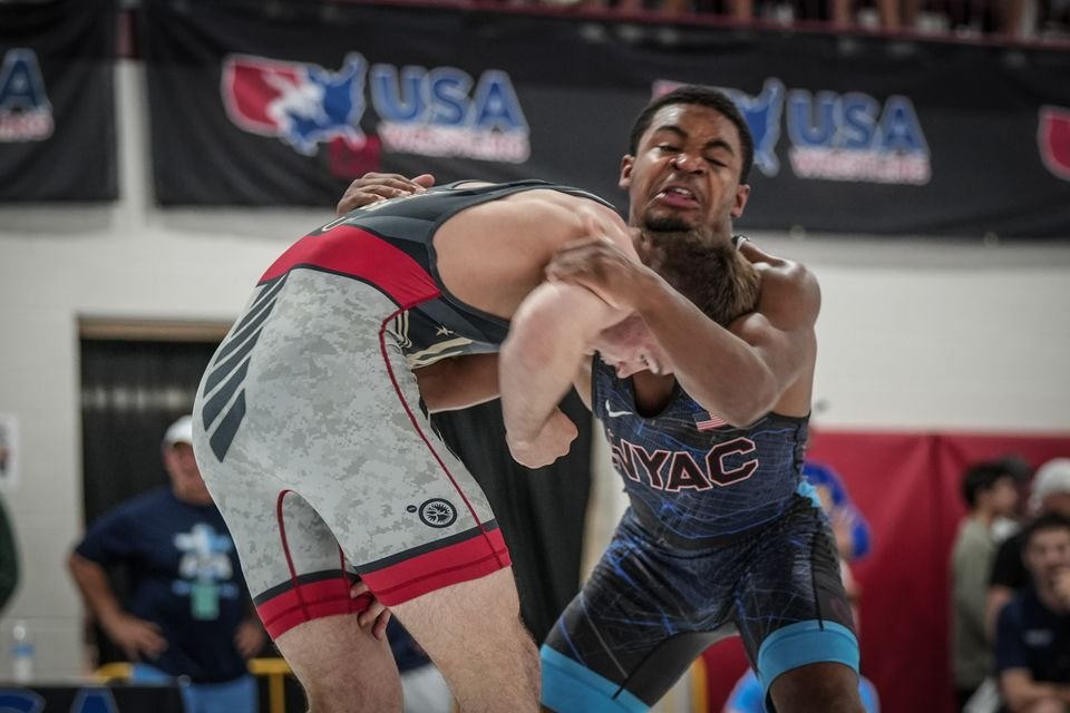 Two wrestlers grapple at close range in front of a USA Wrestling banner.