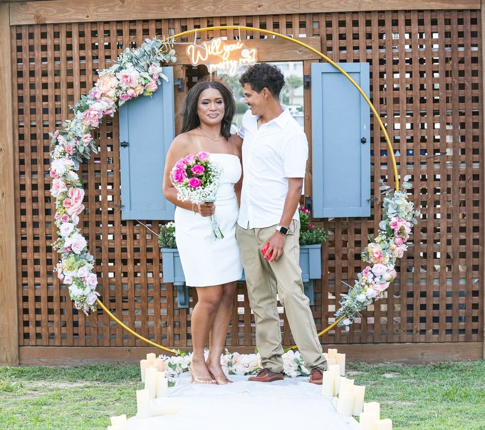 A couple stand together under a floral proposal arch, her holding a bouquet.