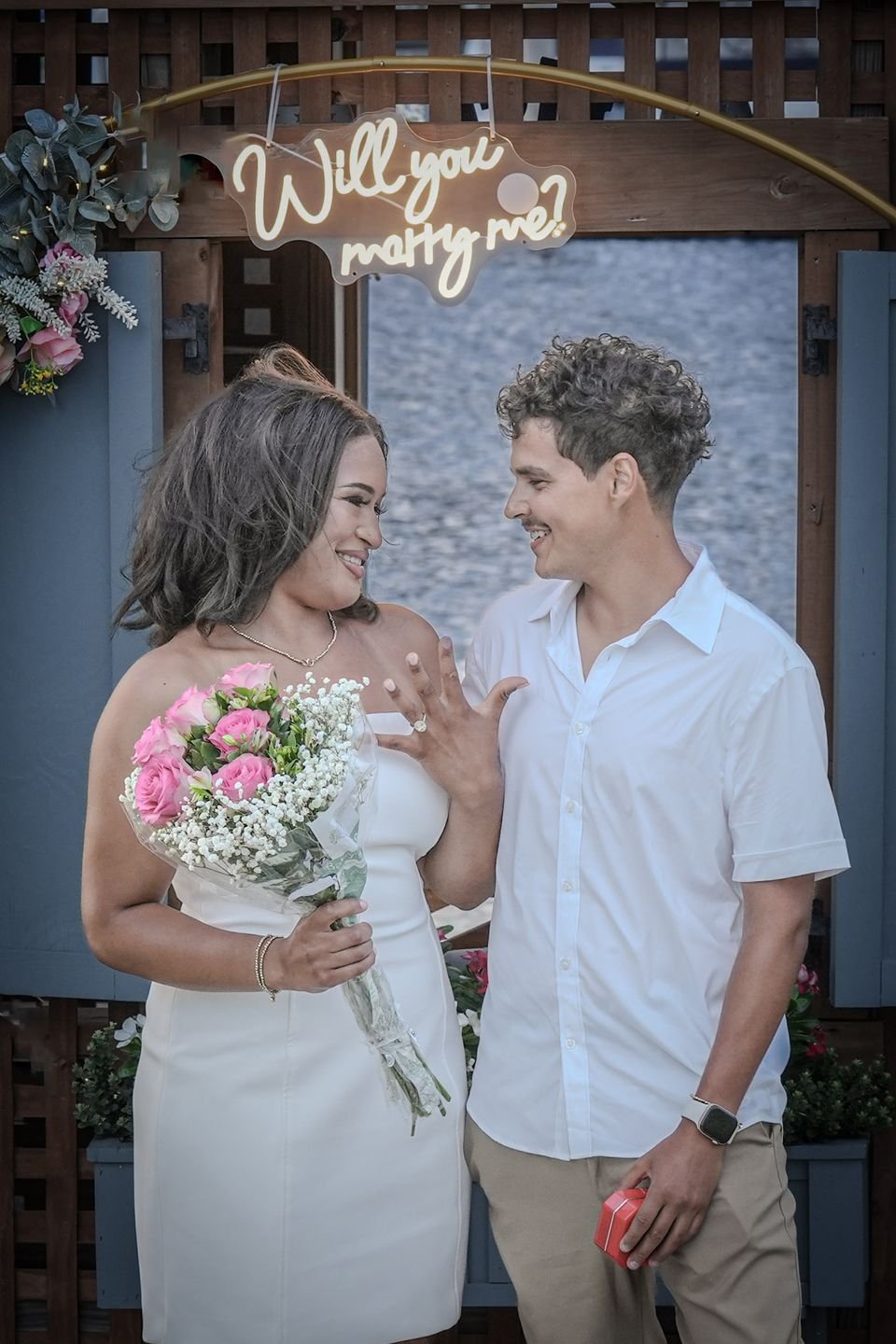 A newly engaged couple smile at each other beside a floral arch, her holding pink roses.