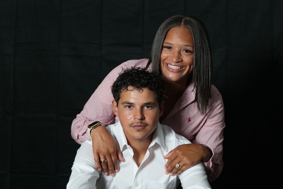 A smiling woman in a pink striped shirt rests her hands on the shoulders of her fiancé, seated in a white shirt, against a black backdrop.