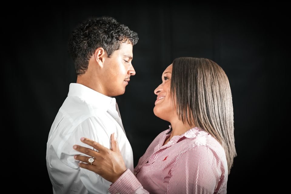 An engaged couple gaze at each other in profile against a black backdrop, her ring hand resting on his chest.