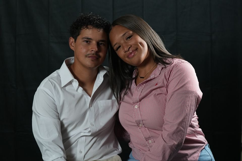 An engaged couple lean together, smiling, against a black studio backdrop.