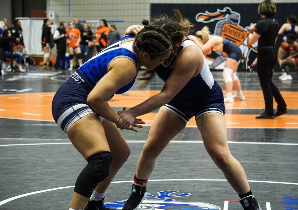 Two women wrestlers in navy and blue tie up on a black and orange mat.