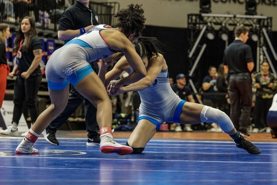 Two women wrestlers in grey and light blue scramble low on a blue mat.