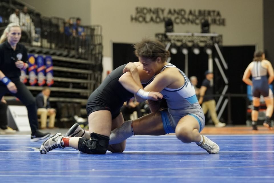 A wrestler in light blue shoots in on her opponent in black on a blue mat.