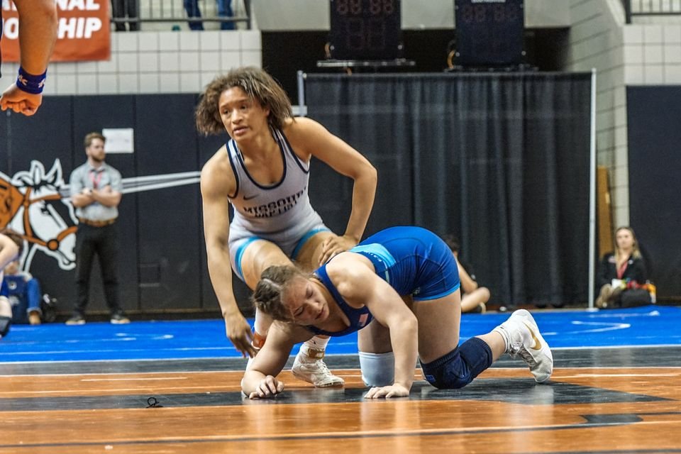 A wrestler in a white singlet works on top of her opponent in blue.