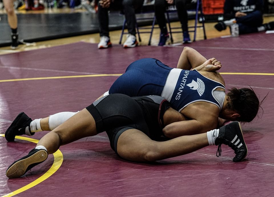 Two women wrestlers scramble on a maroon mat, one driving for control.