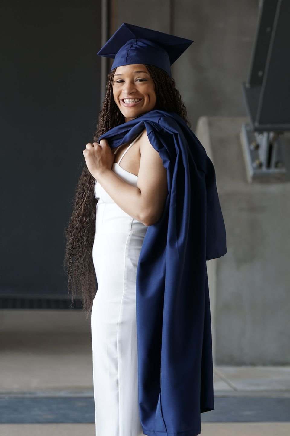 A graduate in a navy cap holds her gown over one shoulder in a white jumpsuit.