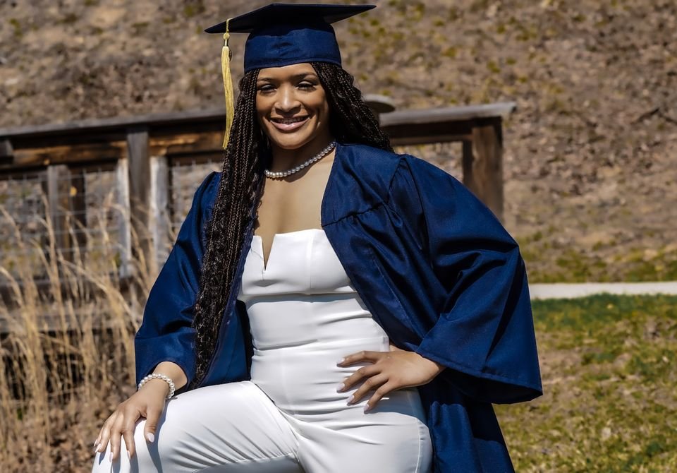 A graduate in a navy cap and gown over a white jumpsuit sits outdoors, smiling.
