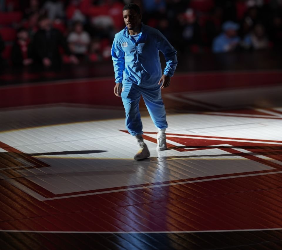 A collegiate wrestler in a Carolina blue warm-up walks out through a single spotlight onto a dark arena floor.
